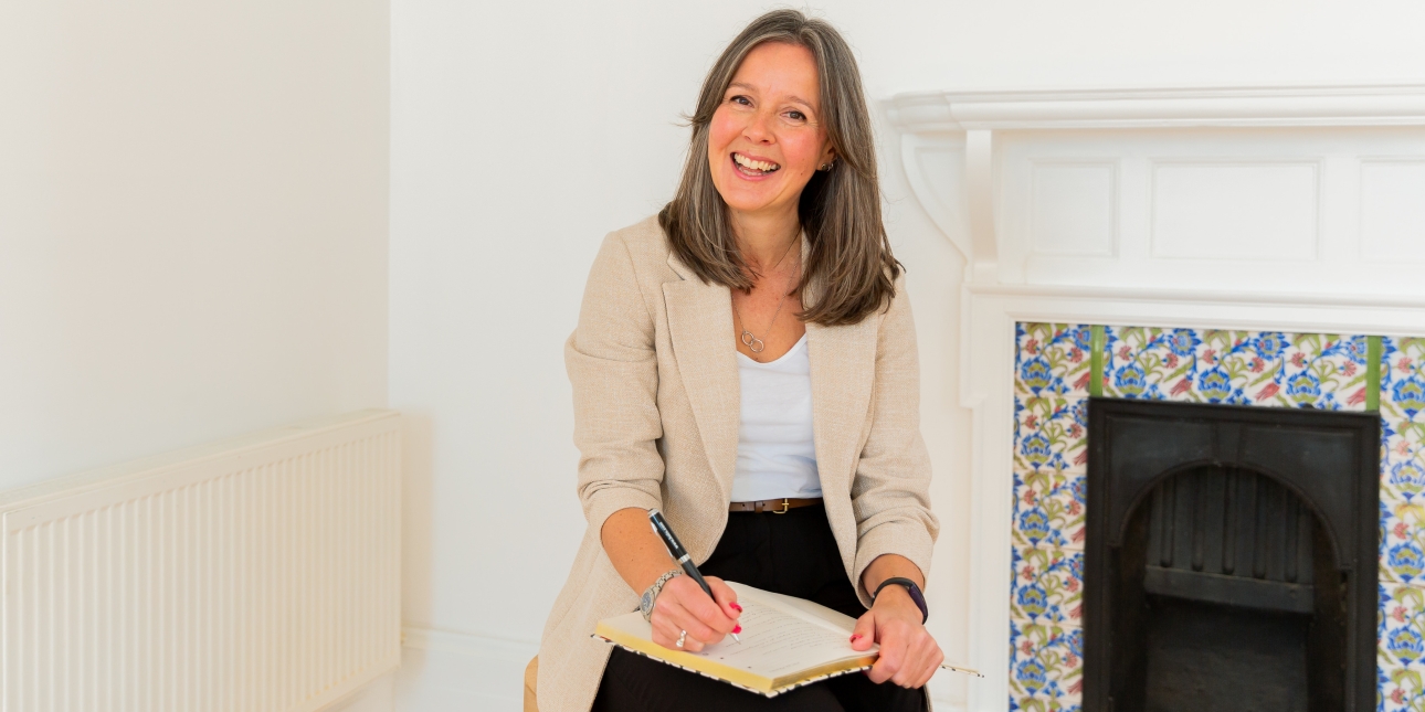 Gemma Pettman, a white woman with shoulder length dark blonde hair sits on a chair in a white room with a tiled fireplace in the background. She is wearing black trousers, a white top and cream jacket and has a notepad on her lap.